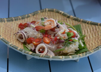 Seafood vermicelli salad served on woven bamboo plate placed on blue wooden floor. Spicy dish consisting of glass noodles, squid,shrimp, mussels,paprika, tomatoes, seasoned with sour-sweet-salty sauce