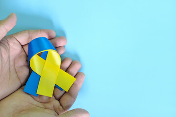 Human hands holding blue and yellow bicolor ribbon on blue background. World down syndrome day, awareness, help and support concept.