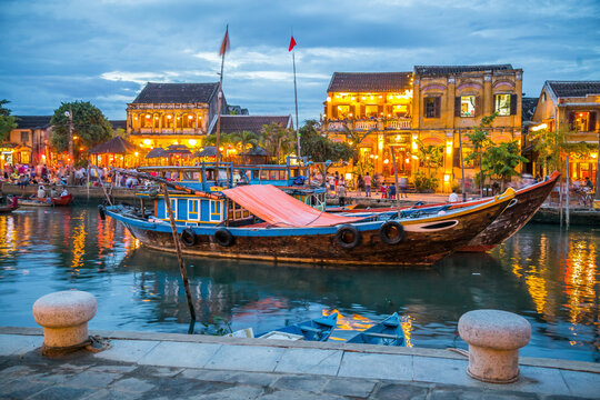 A wooden river boat sitting in front of historic historic buildings lit up at twilight at Hoi An in Vietnam