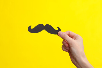 Paper-cut mustache in a female hand on a yellow background. Father's day