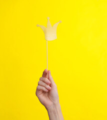 Hand holds paper cut golden crown with stick on yellow background. Party concept