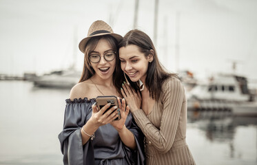 Two surprised cute women friend look at the smartphone screen in the yacht club