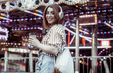 Obraz premium Cute stylish woman in glasses and denim overalls, listening to music on headphones, holding coffee cup in an amusement park
