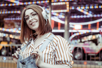 Cute smiling woman in denim overalls and glasses listens to music on headphones in an amusement park