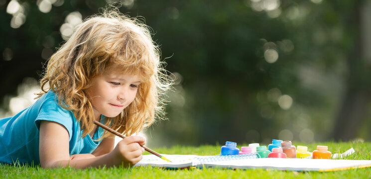 Close up face of cute child outdoors. Spring banner for website header. Artist kids. Kid draws in park laying in grass having fun on nature background.