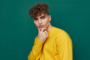 Fototapeta na wymiar a cute, thoughtful man stands on a green background in a yellow sweater holding his hand to the will of the face. Horizontal photo with empty space