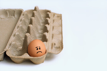 Single sad face chicken egg in an open light brown cardboard dozen egg carton clean white surface...