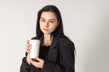 A cute brunette girl holds a cup of coffee in her hands on a white background. A young girl in a black formal business suit is tasting coffee
