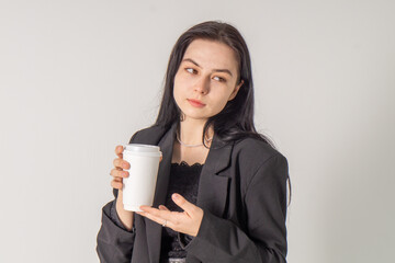 A cute brunette girl holds a cup of coffee in her hands on a white background. A young girl in a black formal business suit is tasting coffee