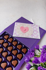 Violet box of heart shaped chocolates, postcard and flowers on white background. Valentines day 14 February sweet present
