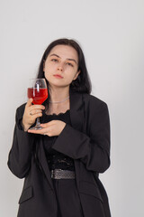 A beautiful young brunette girl drinks red wine, she is deeply unhappy, her heart is broken. Lonely pretty woman dressed in a black business suit, she sits and drinks on a white background