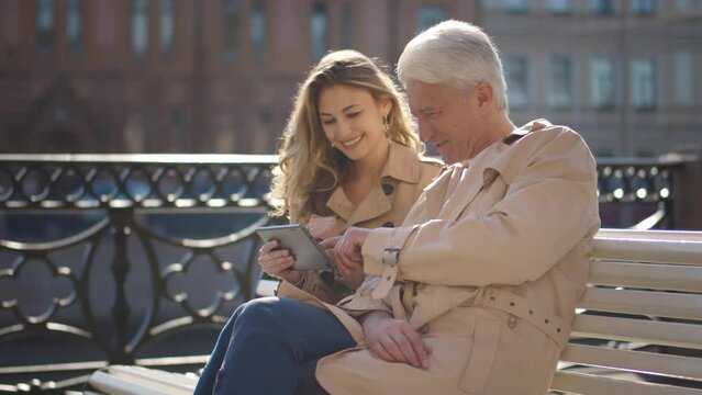 Young daughter show photos on tablet to mature father outdoors. Realtime
