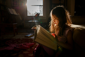 A small child sits in patch of bright sunlight reading a large book
