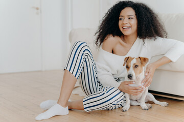 Cute cheerful young African American female sits on floor near sofa, plays with dog, dressed in stylish clothing, looks gladfully somewhere, spends free time at home. Happy pet owner indoor.