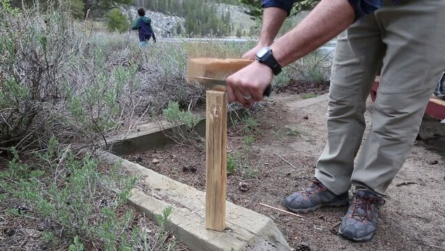 Handheld shot of man chiseling wood while son and dog walking in background at campsite