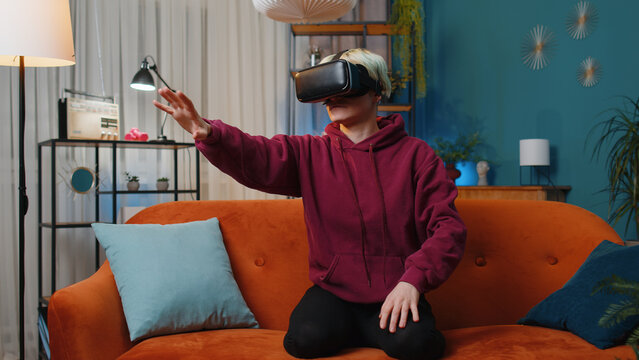 Young woman using virtual reality futuristic technology VR app headset helmet to play simulation 3D 360 video game, watching film movie at modern home apartment. Blonde girl in goggles sitting on sofa - Powered by Adobe