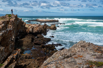 A man standing alone on rocky cliffs along the Cliff Path looking out to Walker Bay and the ocean waves. Hermanus, Whale Coast, Overberg, Western Cape, South Africa.