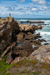 Fototapeta na wymiar A man standing alone on rocky cliffs along the Cliff Path looking out to Walker Bay and the ocean waves. Hermanus, Whale Coast, Overberg, Western Cape, South Africa.