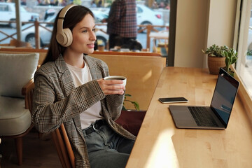 Young woman with thoughtful facial expression listening to a podcast on a laptop. Female freelancer in headphones having a coffee break from working in a coffeehouse. Close up, copy space, background.