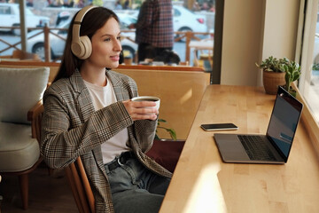 Young woman with thoughtful facial expression listening to a podcast on a laptop. Female freelancer...