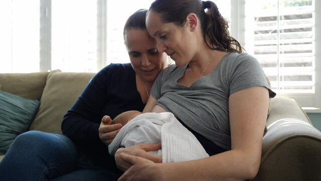 Handheld shot of mother breastfeeding son while sitting with woman on sofa at home