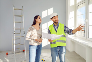 Male builder talking to woman. Happy, smiling foreman in hardhat and uniform vest standing in new...
