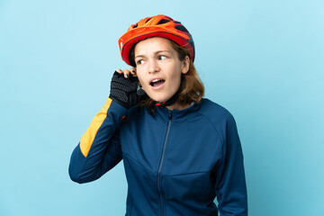 Young cyclist woman isolated on blue background listening to something by putting hand on the ear