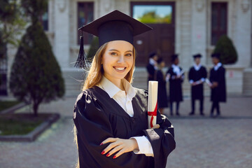 Young attractive Caucasian woman posing smilingly for student portrait holding certificate of higher education grant wearing black academic gown and hat standing in front of college building - obrazy, fototapety, plakaty
