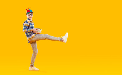 Happy school child having fun. Side view full length cheerful boy in casual shirt, jeans, funny propeller cap and round eyeglasses standing on yellow copy space background, holding books and smiling