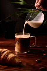 hand pours milk into a glass cup with coffee, which stands on a wooden table with a croissant,...