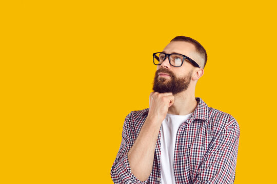 Portrait of an intelligent bearded young man in a casual shirt and glasses holding his hand on chin, thinking about something and looking away at the solid yellow color copy space background