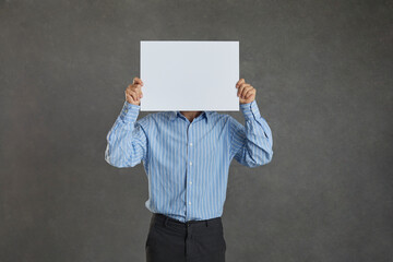 Studio portrait of unrecognizable young man covering his face hiding behind blank mockup banner. Anonymous businessman holding clean empty white sheet of paper standing isolated on grey background