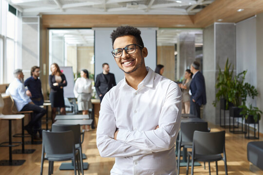Portrait of happy Afro American business coach. Handsome young Black man in white shirt and eyeglasses standing in office after corporate team training class, looking at camera and smiling