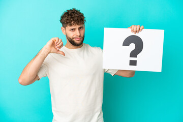 Young handsome caucasian man isolated on blue background holding a placard with question mark symbol and doing bad signal