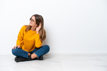 Young caucasian woman sitting on the floor isolated on white background listening to something by putting hand on the ear