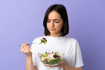 Young caucasian woman isolated on purple background holding a bowl of salad with sad expression