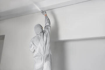 Decorator in protective overalls painting ceiling with spray gun indoors