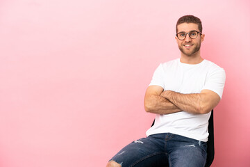 Young man sitting on a chair over isolated pink background keeping the arms crossed in frontal...