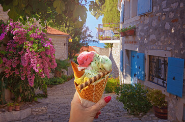 Woman hand hold  beautiful bright sweet ice - cream cone with different flavors  held in hand on the background of old street  in  Rovinj .Rovinj is a tourist destination on Adriatic coast of Croatia - 568010588