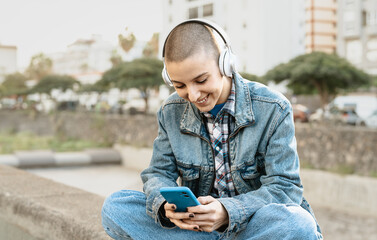 Shaved head girl using mobile smartphone while listening to music with headphones in the city street