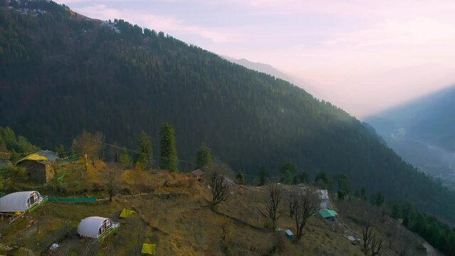 aerial drone shot panning while focussed on geodesic and triangular tents on side of himalaya to sun setting behind mountain showing tourists doing glamping in hamta village manali