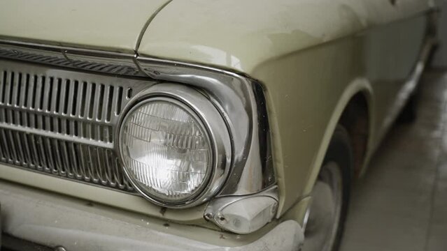 Vintage beige car with round headlights and crome details in collectible condition under layer of dust in tiled room. On chrome parts, reflections of moving people are visible.