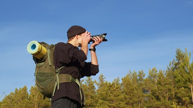 Handsome guy with professional camera takes pictures in woods, evaluates result. Male tourist with backpack, mat is shooting of forest landscapes. Cameraman working on nature. Side view