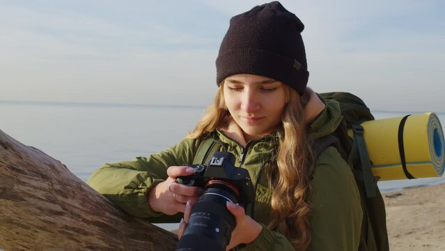 Pretty long-haired girl looks to camera at photographs taken, smiles near river bank in autumn. Young lady portrait in beanie, jacket standing on sandy beach near dry log seeing at pictures on screen