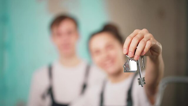 Happy, young, in love guy and girl rejoice in moving to new apartment and beginning of renovation. Holds in hands key to apartment with keychain house. Start make repairs. Married couple laugh, smile.