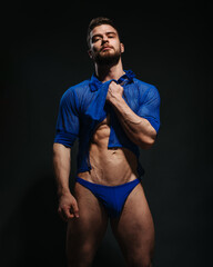 Sexy muscled male model in blue shirt and blue swimwear on black background. Bearded man taking his clothes up. Fashion guy in beachwear in studio. Male fitness model in stylish outlook. - obrazy, fototapety, plakaty