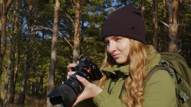 Handsome girl with professional camera takes pictures in woods, evaluates result. Female tourist with backpack is shooting of forest landscapes. Cameraman working on nature