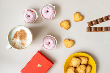 on a beige background, a mug of cappuccino, a red box with a gift, candles, heart-shaped cookies, pink marshmallows, chocolate