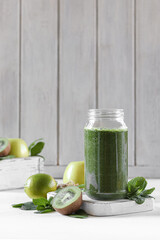A green smoothie in a glass jar stands on a white wooden background, next to greens, vegetables and fruits.