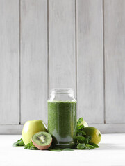 A green smoothie in a glass jar stands on a white wooden background, next to greens, vegetables and fruits. Healthy food.